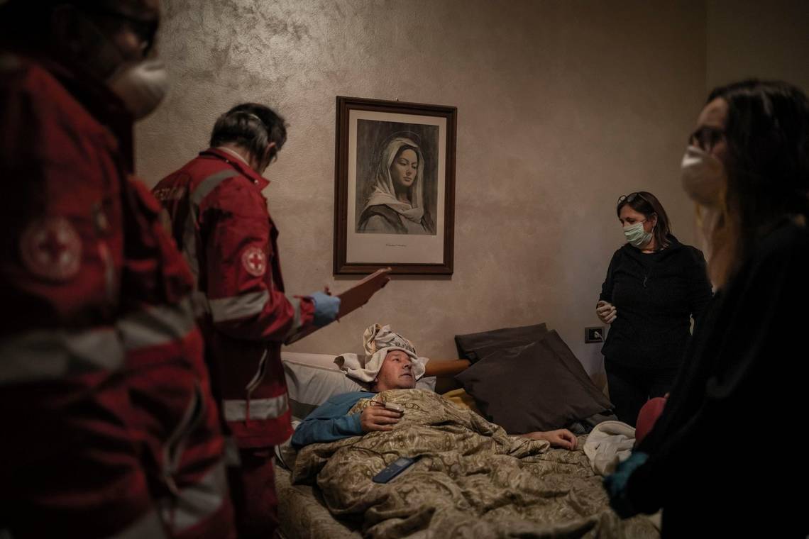 A man lies in bed surrounded by Italian Red Cross volunteers. A painting of the Virgin Mary hangs on the wall. Photo by Fabio Bucciarelli.