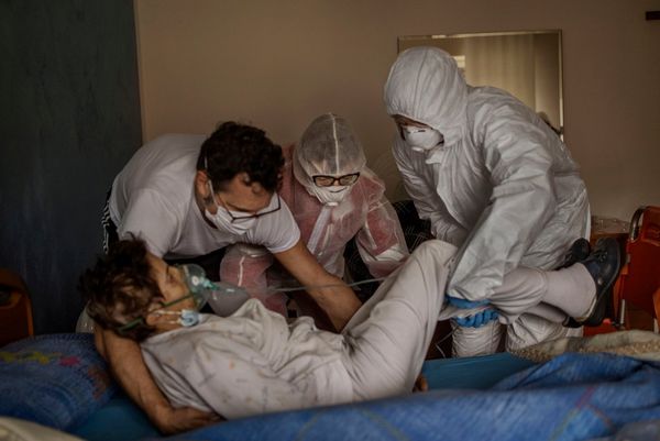 Three people in protective gear help lift a woman with Covid-19 onto her bed. Photo by Fabio Bucciarelli.
