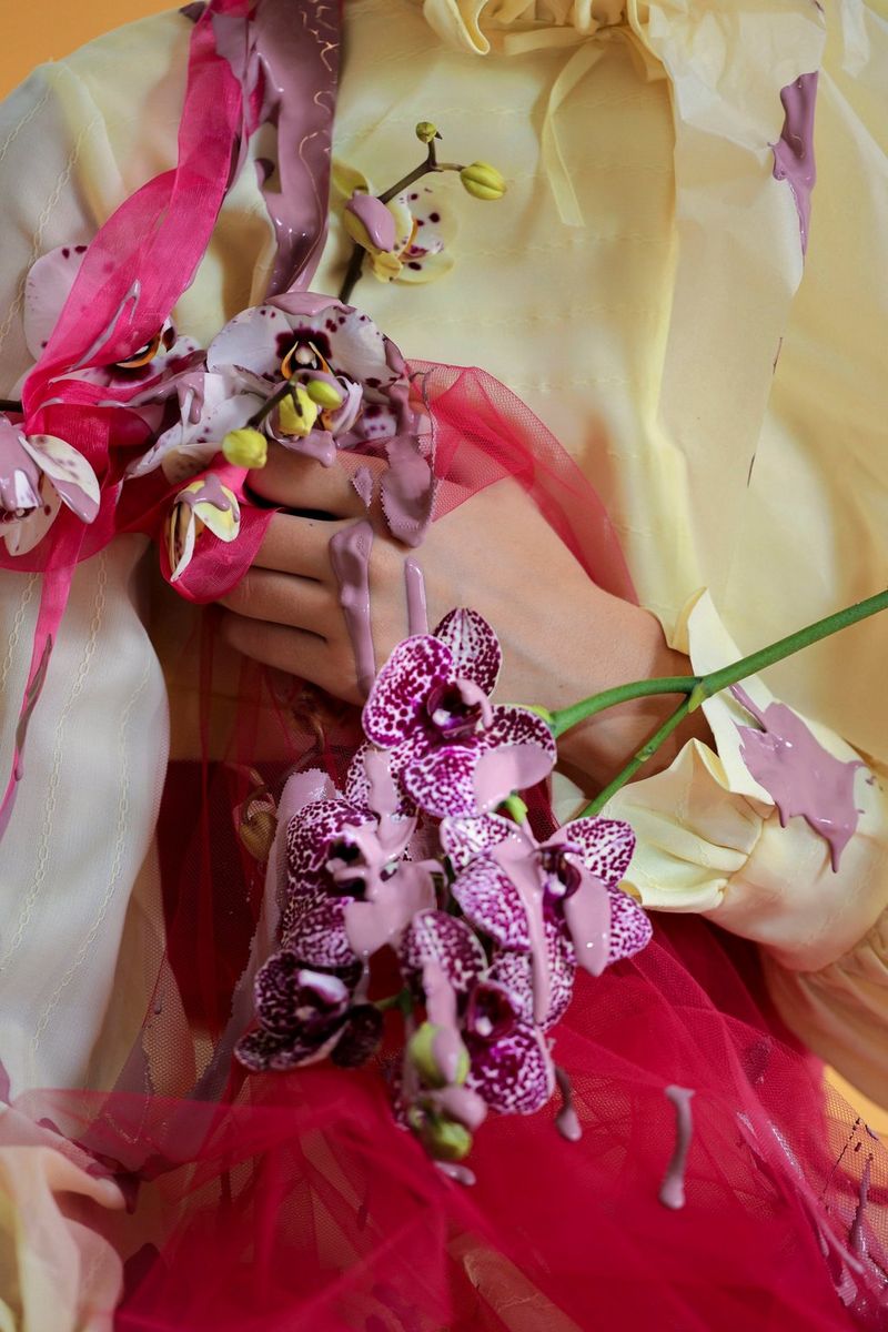 A close-up of a model wearing an abstract outfit, adorned by flowers with purple paint poured on her.