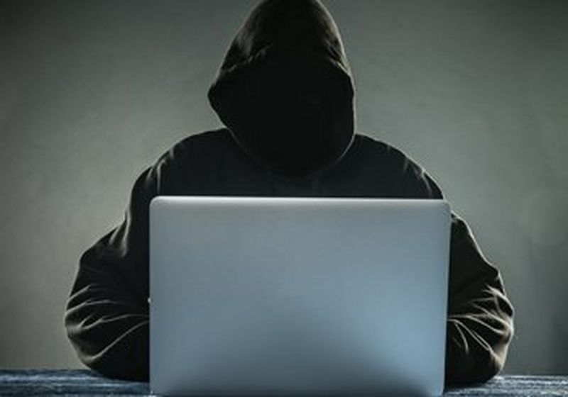 A hooded figure sits behind a laptop.