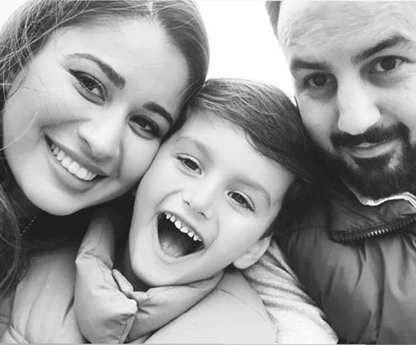 A black and white photo of a woman, a young child and a man hugging together for a selfie. They wear winter coats and are all smiling and happy.