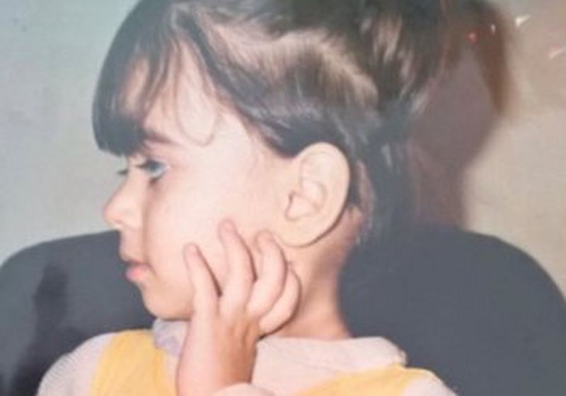 A head and shoulders photo of a little child, wearing a white and yellow top and with a high ponytail, held with a red bow. She is facing left and has one hand raised to her cheek.
