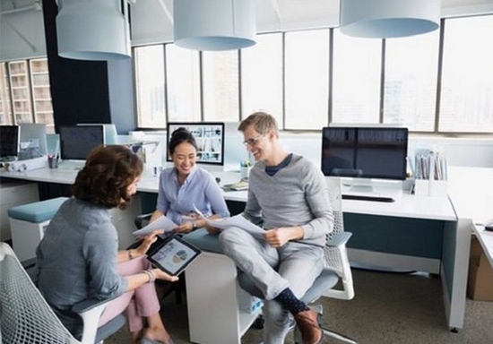 Two women and a man sitting in front of a long white desk adorned with PCs compare notes between pieces of paper and a tablet device.