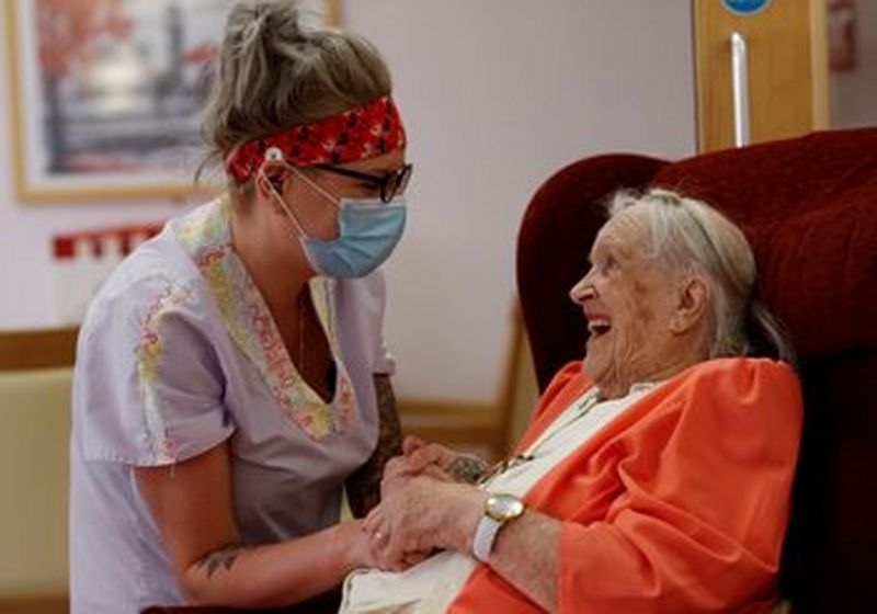 A woman in a medical mask and overalls holds the hands of a smiling elderly woman who wears an orange cardigan.