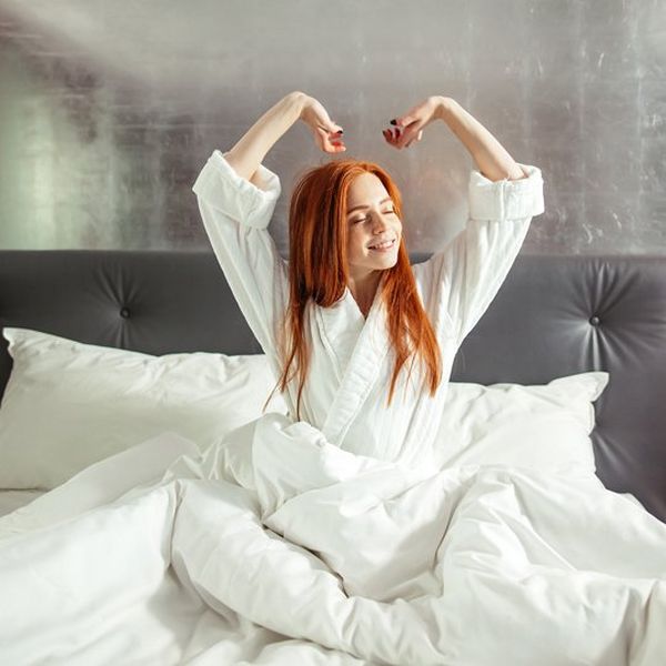 A young woman with red hair sits up in bed and stretches. The bed clothes are soft and white, the headboard grey and she wears a white dressing gown.