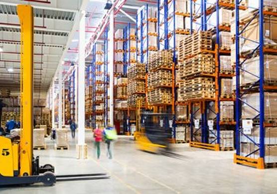 An action shot of a warehouse, with forklift trucks and people moving between huge racks of boxes on pallets, awaiting shipment.
