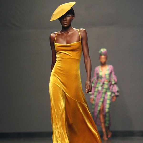 A model on a catwalk wearing a mustard yellow slip dress and hat
