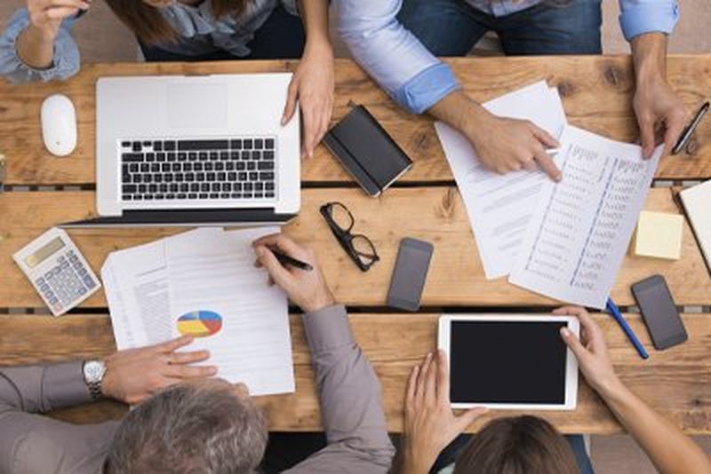 Two women perch on long white desks alongside computers as they chat to three colleagues sitting in front of them.