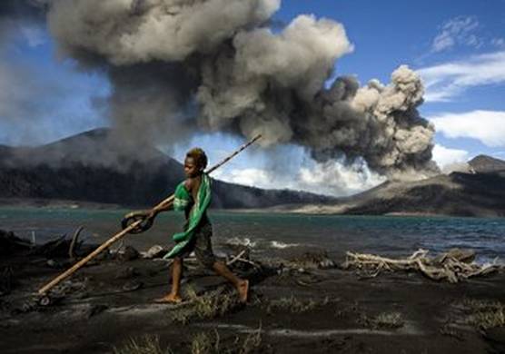 A boy stands, playing, on a grey ash beach by the sea, in the background a volcano is erupting against a blue sky. © Ulla Lohmann