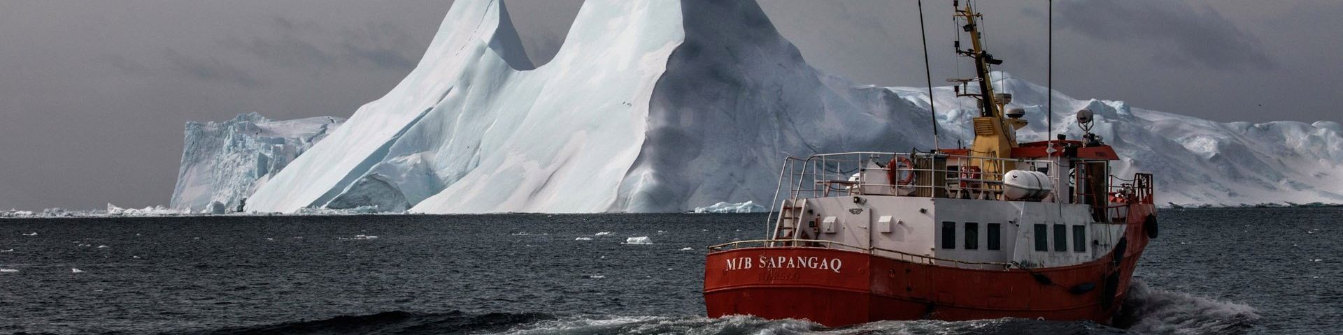 A red boat sails on cold grey water, with a huge floating iceberg behind it.