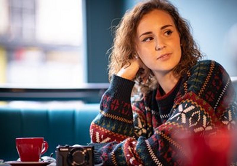 A young woman sits in a coffee shop, head resting on her hand, looking into the distance. She wears a colourful sweater.
