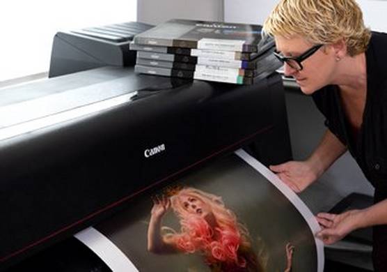Cheryl stands to the right, with short, blonde hair and black framed glasses. She gently holds a print as it emerges from a Canon PRO-4000 printer.