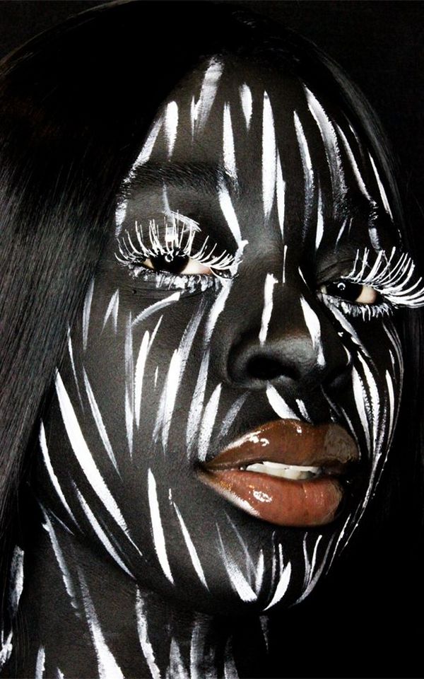 A woman's face, painted black with white brushstrokes. Her lips are high gloss and eyelashes painted white.