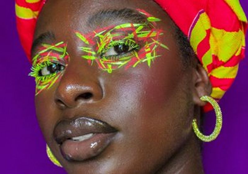 The face of a woman as she looks directly at the camera. She wears a painted neon yellow and red crosshatch pattern on her eyes, a matching head-wrap and neon yellow hoop earrings. © Wendy Asumadu