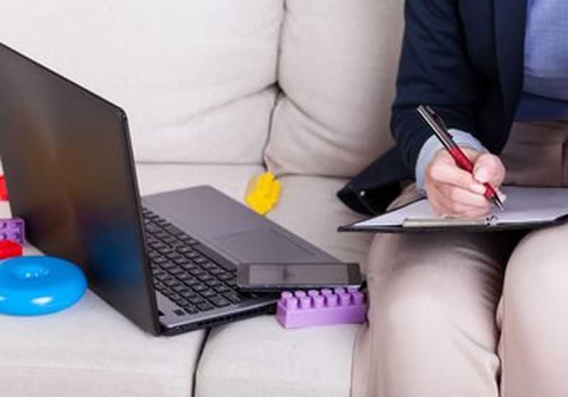 On the right, the lower body of a person sat on a couch and wearing beige trousers. They have a clipboard on their lap and is writing with a red pen. Beside them on the left is an open grey laptop and a mobile phone. They are surrounded by children’s colourful building blocks.