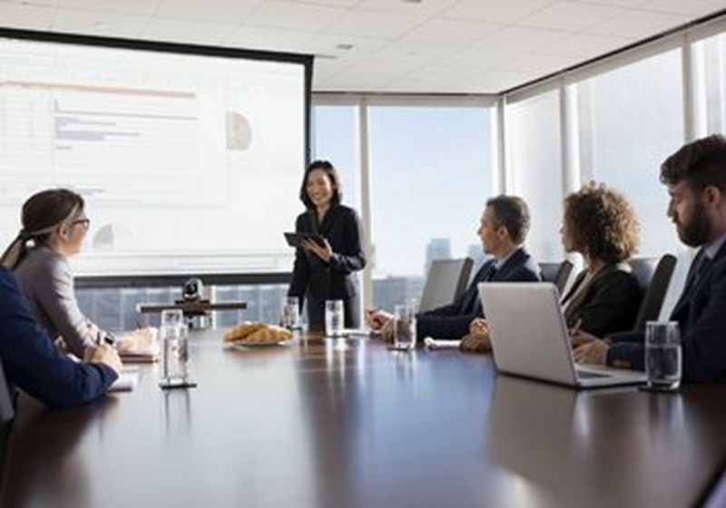 A woman holding a tablet gives a presentation in front of a projector screen for five colleagues, who are sat at a boardroom table with laptops, glasses of water and a plate of croissants.