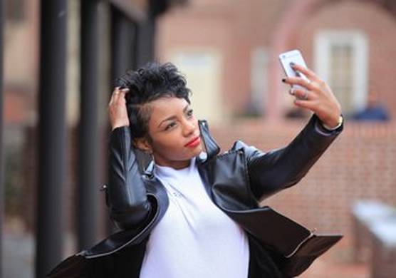 A young woman in a white t-shirt and leather jacket, adjusts her hair as she takes a selfie.