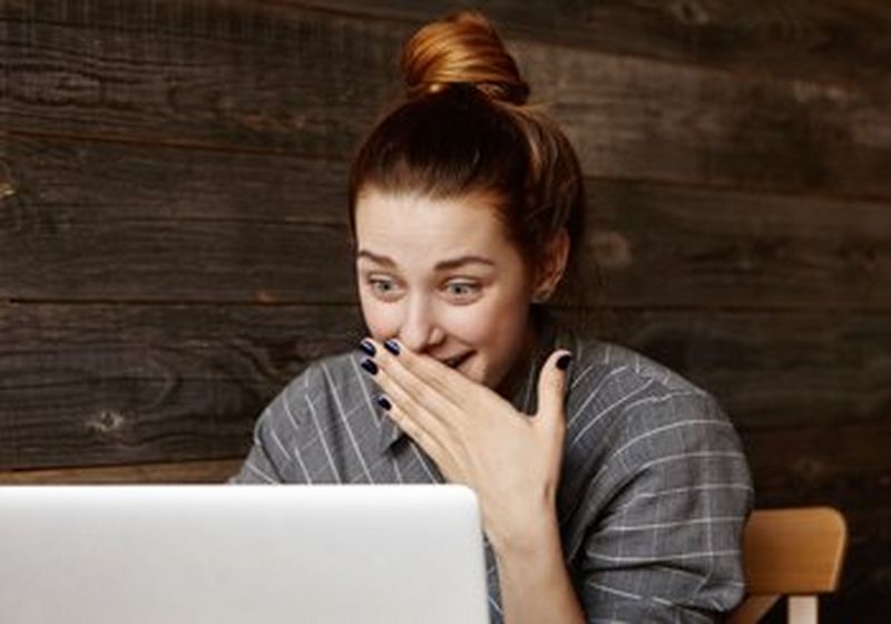 A woman with her red hair in a bun, sits in front of a laptop. She wears a grey and white checked shirt. The background is dark wood. She looks shocked and has her hand covering her mouth in disbelief. Her fingernails are painted black.