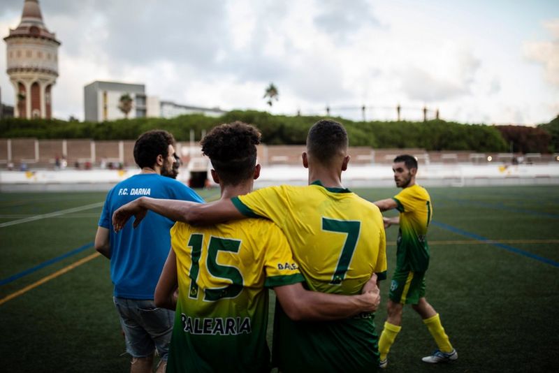 Dos jugadores de fútbol en un campo de césped artificial, de espaldas a la cámara, con los brazos sobre los hombros del otro. Detrás de ellos, a la izquierda, aparece una persona con camiseta azul y pantalones cortos claros de espaldas a la cámara, mientras que, a la derecha, se ve ligeramente difuminado por el movimiento otro jugador con un uniforme similar amarillo y verde. Cortesía del proyecto Goals for Change. Creado por Ofelia de Pablo, Javier Zurita y Pablo Tosco.