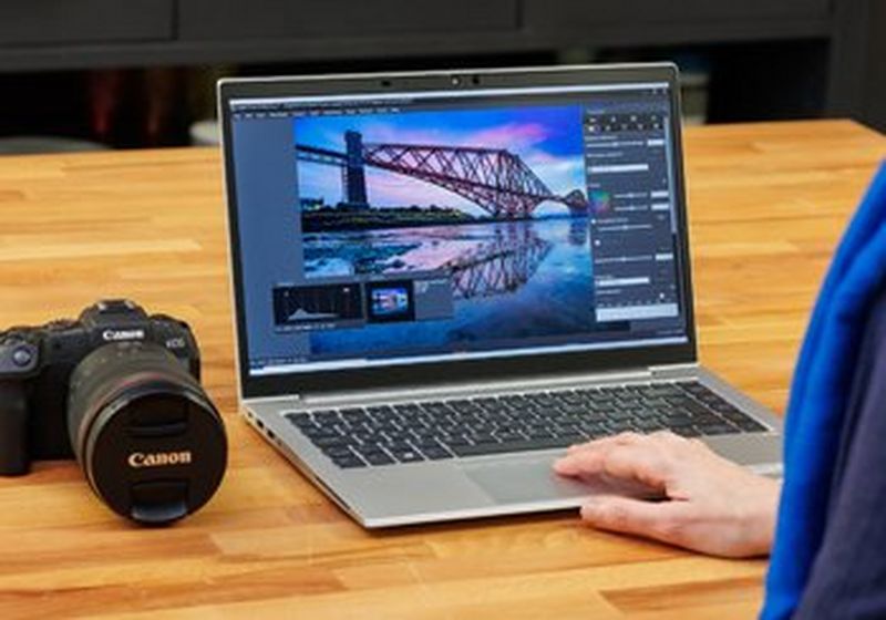 A wooden table with a Canon camera and a laptop sat upon it. The laptop shows a screen with a photo editing application and one hand is on the keyboard.