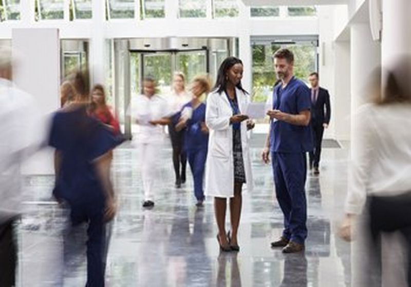 Two doctors stand looking at a document in a busy corridor. Around them are the blurred figures of people moving past them quickly.