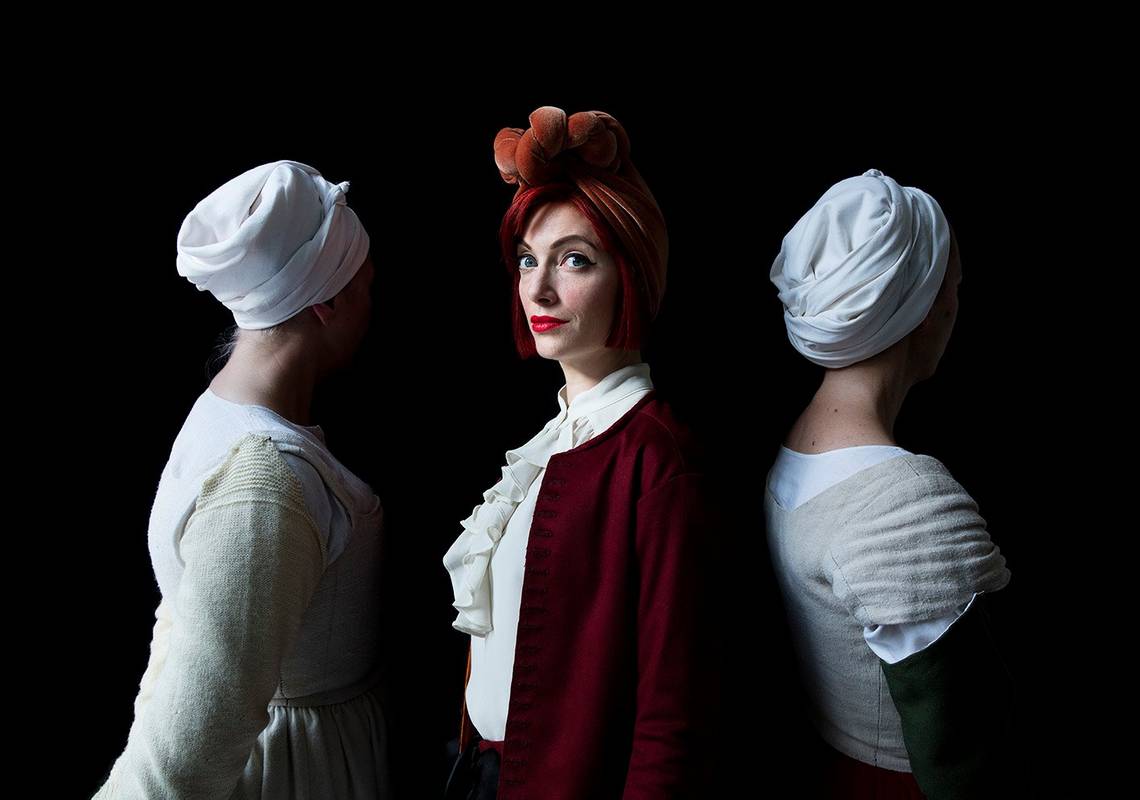 A woman with red hair stands side-on, facing the camera. On either side of her are women facing the other way, wearing old-fashioned white servant-style hats and dresses. Theyre against a black background.