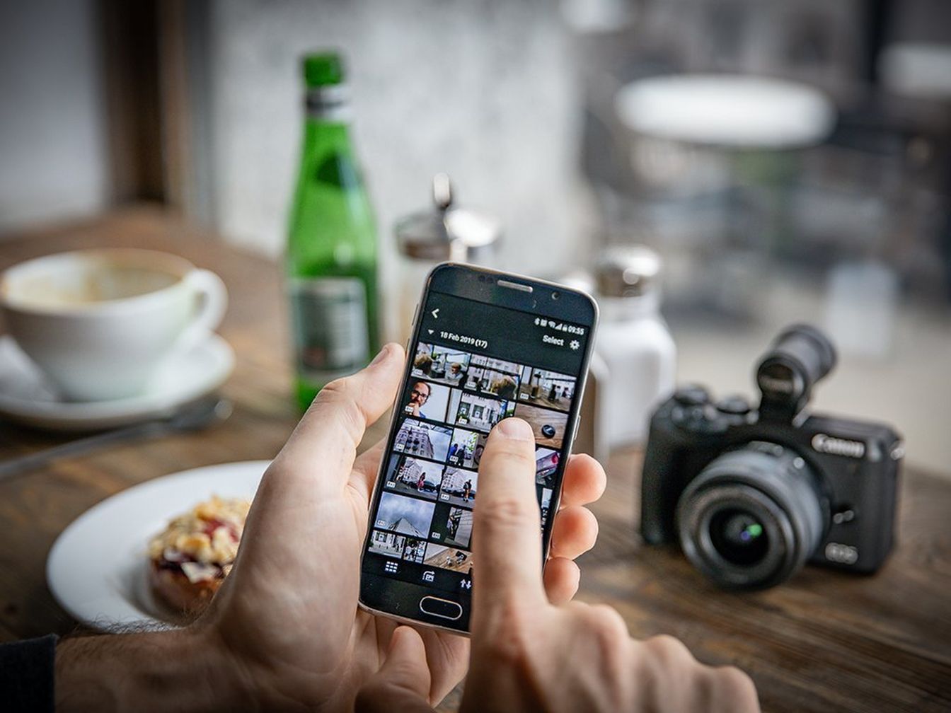 يدان تحملان هاتفًا ذكيًا عليه صور في تطبيق Camera Connect، وتظهر في الخلفية الكاميرا على الطاولة مع الطعام والمشروبات.