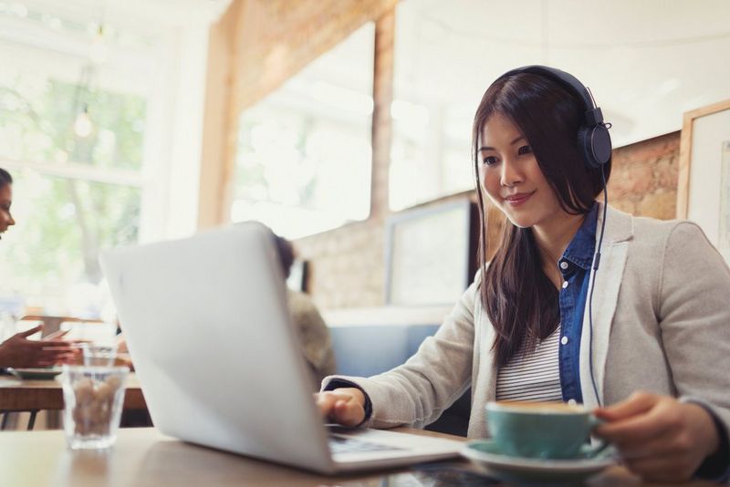 A business woman smiling working on a laptop with headphones on in a cafe.