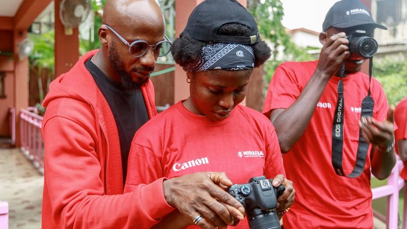 A bespectacled male Canon Certified Trainer stands behind a woman and they both look into the back screen of the camera she holds. To their right is a man holding a Canon camera to his face.
