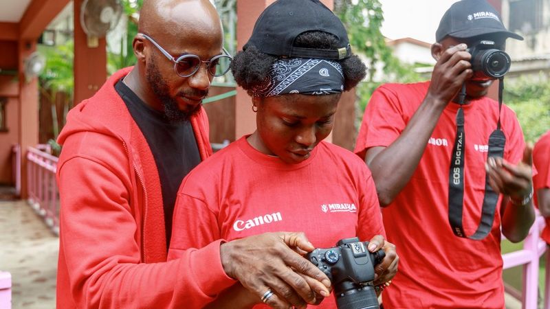 Un formateur certifié Canon portant des lunettes se tient derrière une femme. Ils regardent tous les deux l'écran arrière de l'appareil photo que la femme tient. À leur droite, un homme tient un appareil photo Canon près de son visage.