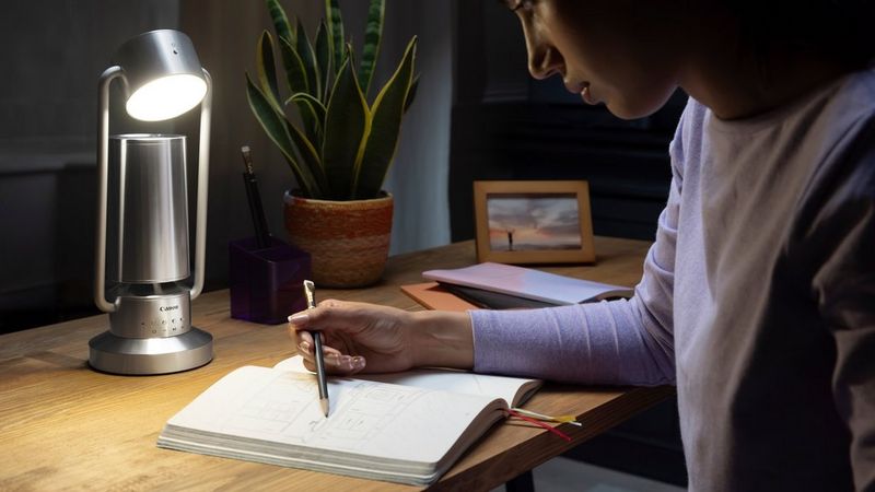 A woman sits at a desk, looking at her notebook and holding a pencil to the page. On the table sits a silver Canon Light & Speaker ML-A, beside a potted plant and a picture in a frame.