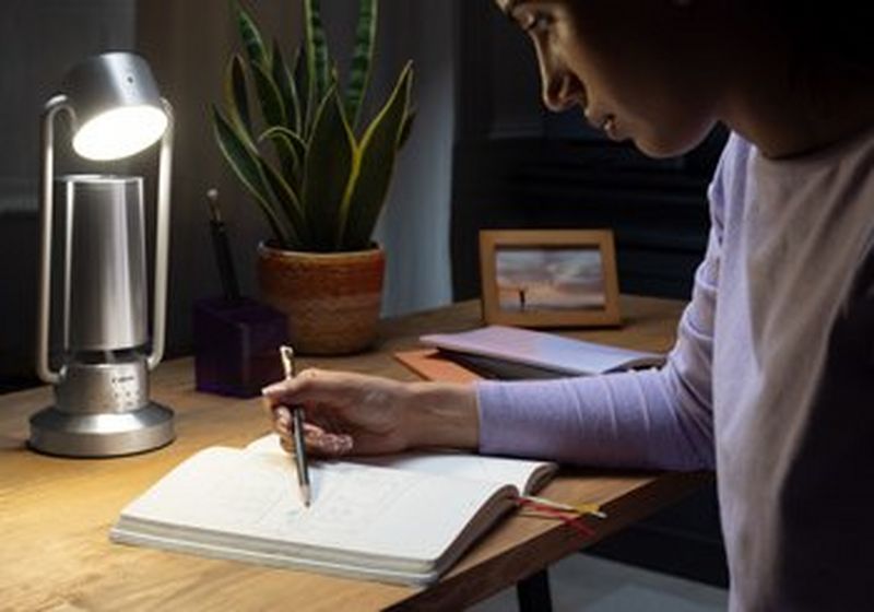 A woman sits at a desk, looking at her notebook and holding a pencil to the page. On the table sits a silver Canon Light & Speaker ML-A, beside a potted plant and a picture in a frame.