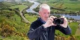 Alan Rowan stands on the side of a hill taking a photo using a Canon compact camera, with a river in the valley behind him.