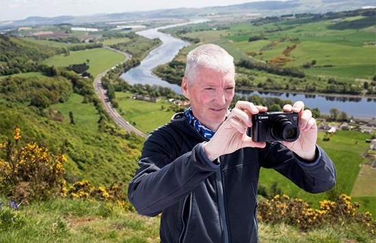 Alan Rowan stands on the side of a hill taking a photo using a Canon compact camera, with a river in the valley behind him.