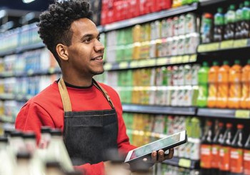 A young man in a uniform red top and black apron stands in front of supermarket shelves, smiling. He holds a tablet computer and looks as though he is a retail worker.