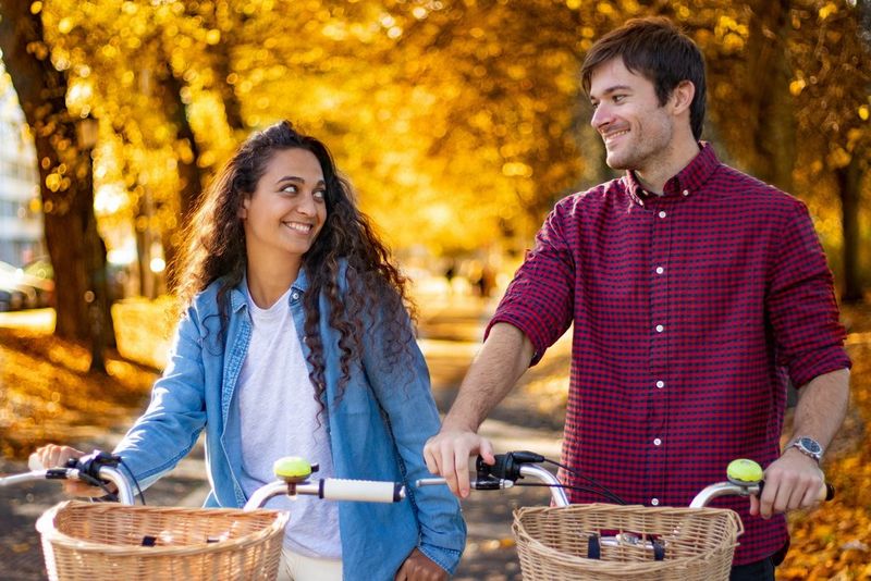 شاب وفتاة يبتسمان إلى بعضهما في أثناء ركوبهما دراجتيهما على طول مسار مقابل خلفية من أوراق الخريف الذهبية.