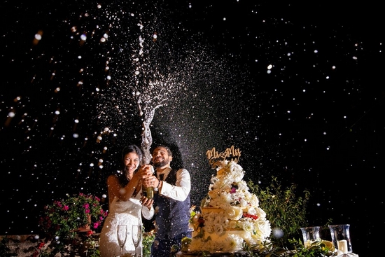 A spurt of champagne frozen in the air as smiling newlyweds celebrate next to a towering cake at their wedding reception. 