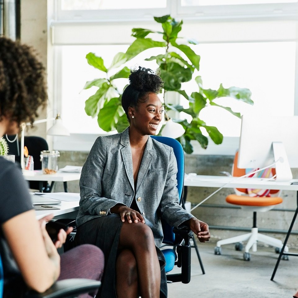 Woman sits on blue chair in front of a plant and talks to two people