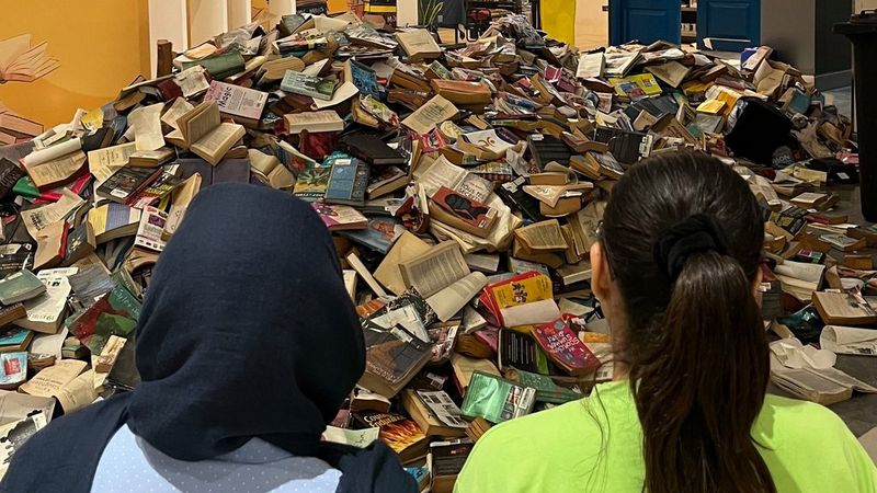 The head and shoulders of two women, photographed from behind, as they look at a mountain of damaged books