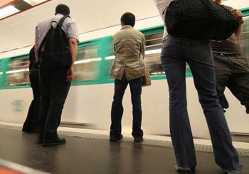 Een foto, genomen vanaf de grond, van vijf personen die op een metroperron staan. Een wit-groene trein rijdt langs.