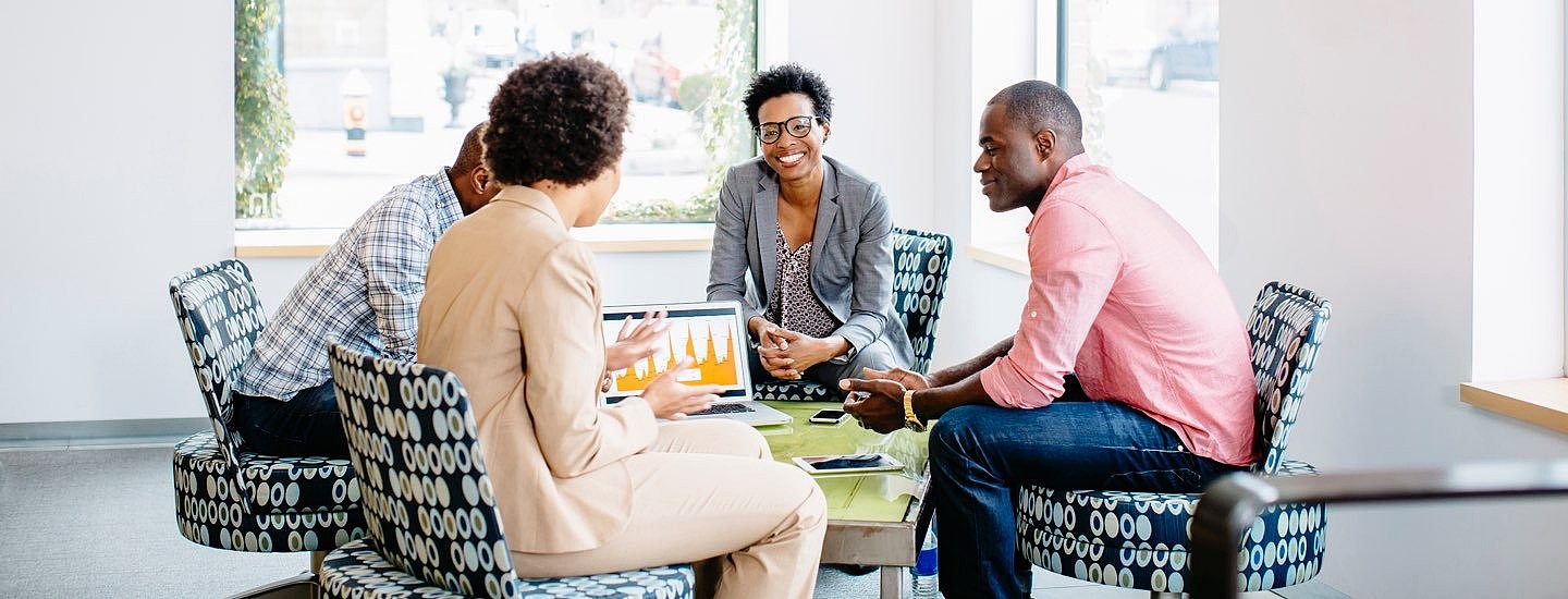 Two men and two women sit round a small table on comfy patterned black and white chairs looking at graphs on a laptop.