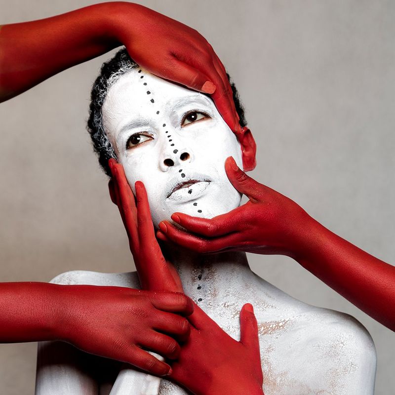 A head-and-shoulders shot of a woman in white body paint, with red painted hands holding her face. Taken by Aïda Muluneh on a Canon EOS 5D Mark III.