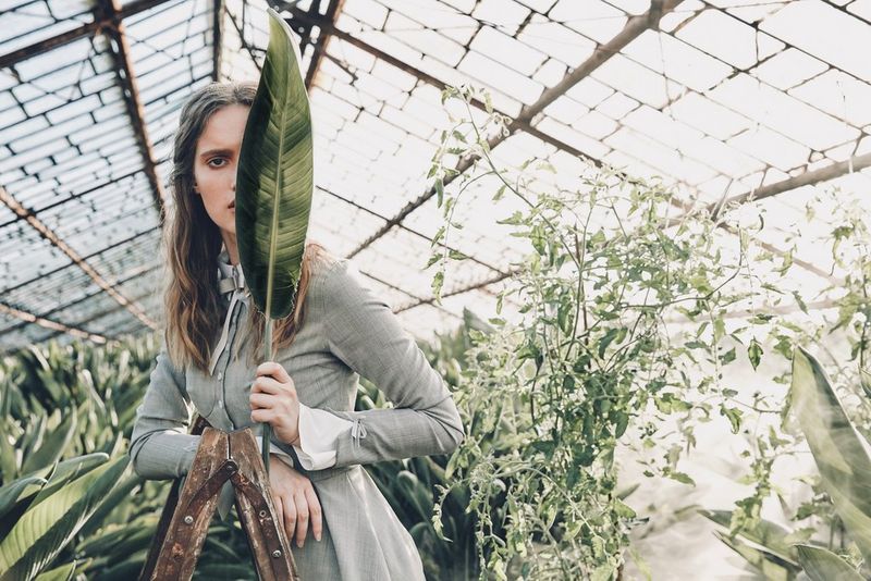 A woman stands in a greenhouse peering out from behind a large leaf. Taken by Jaroslav Monchak on a Canon EOS 5DS R.