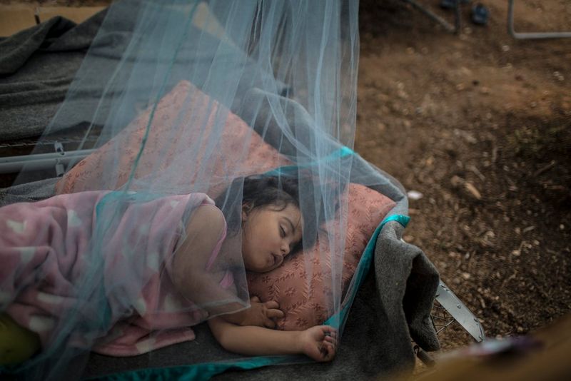 In a refugee camp north of Athens, a young refugee girl sleeps outside under a mosquito net to escape the heat in her family's tent, photographed by Muhammed Muheisen on a Canon EOS 5D Mark III.