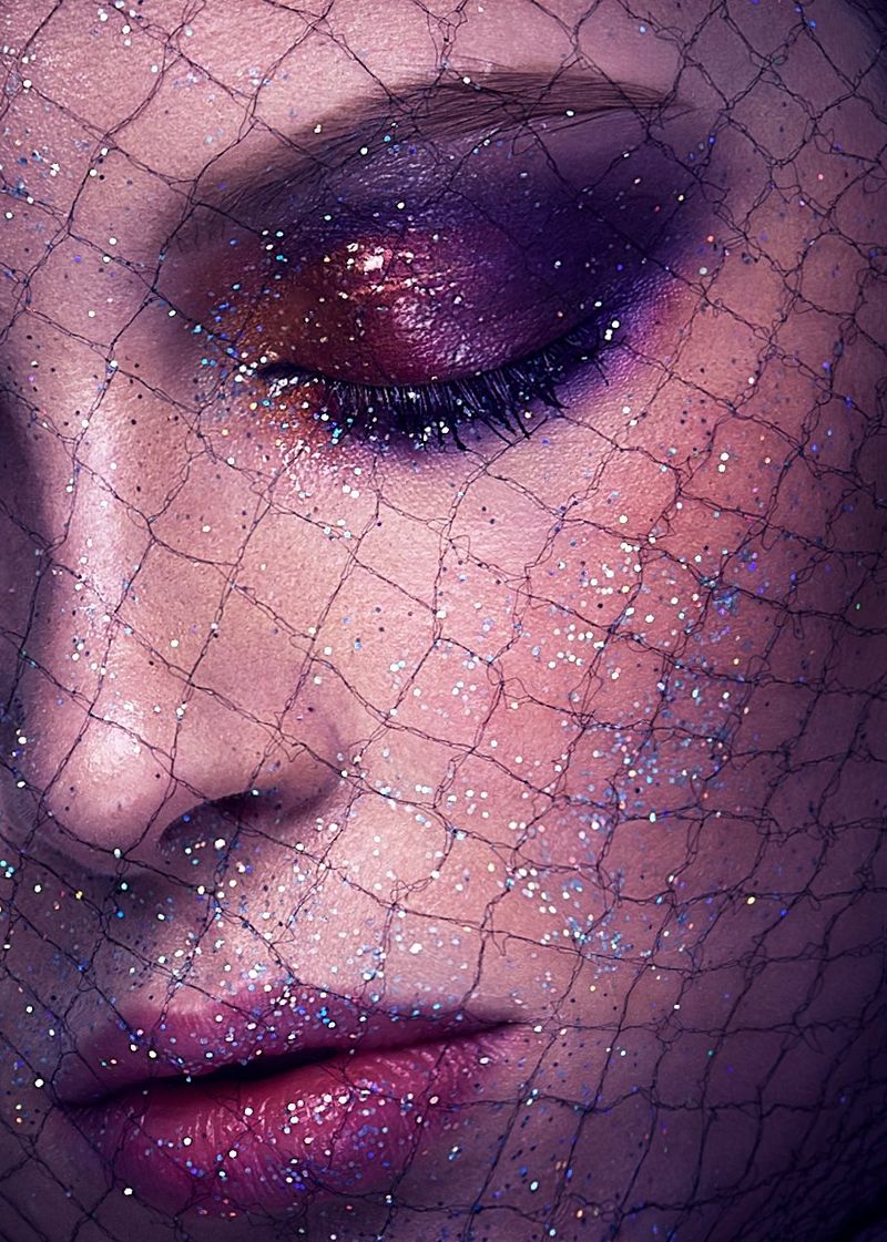 A close up of a woman's face, with bold purple makeup and glitter, and fishnet fabric on her face. Taken on a Canon EOS-1D X by Canon Ambassador Quentin Caffier.