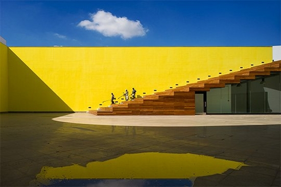 A bright yellow wall with brown stairs leading into a sunny courtyard, with glass doors beneath the stairs. Photo by Fernando Guerra with a Canon TS-E17mm f/4L lens.
