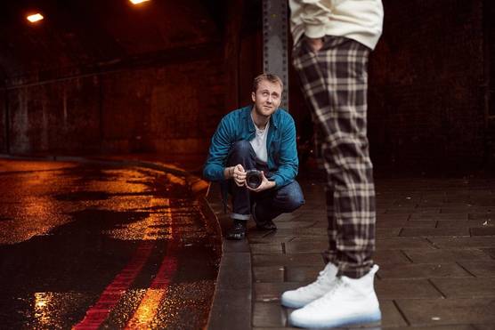 A young man in casual dress crouching on a pavement photographing a man in checkered trousers and white trainers.