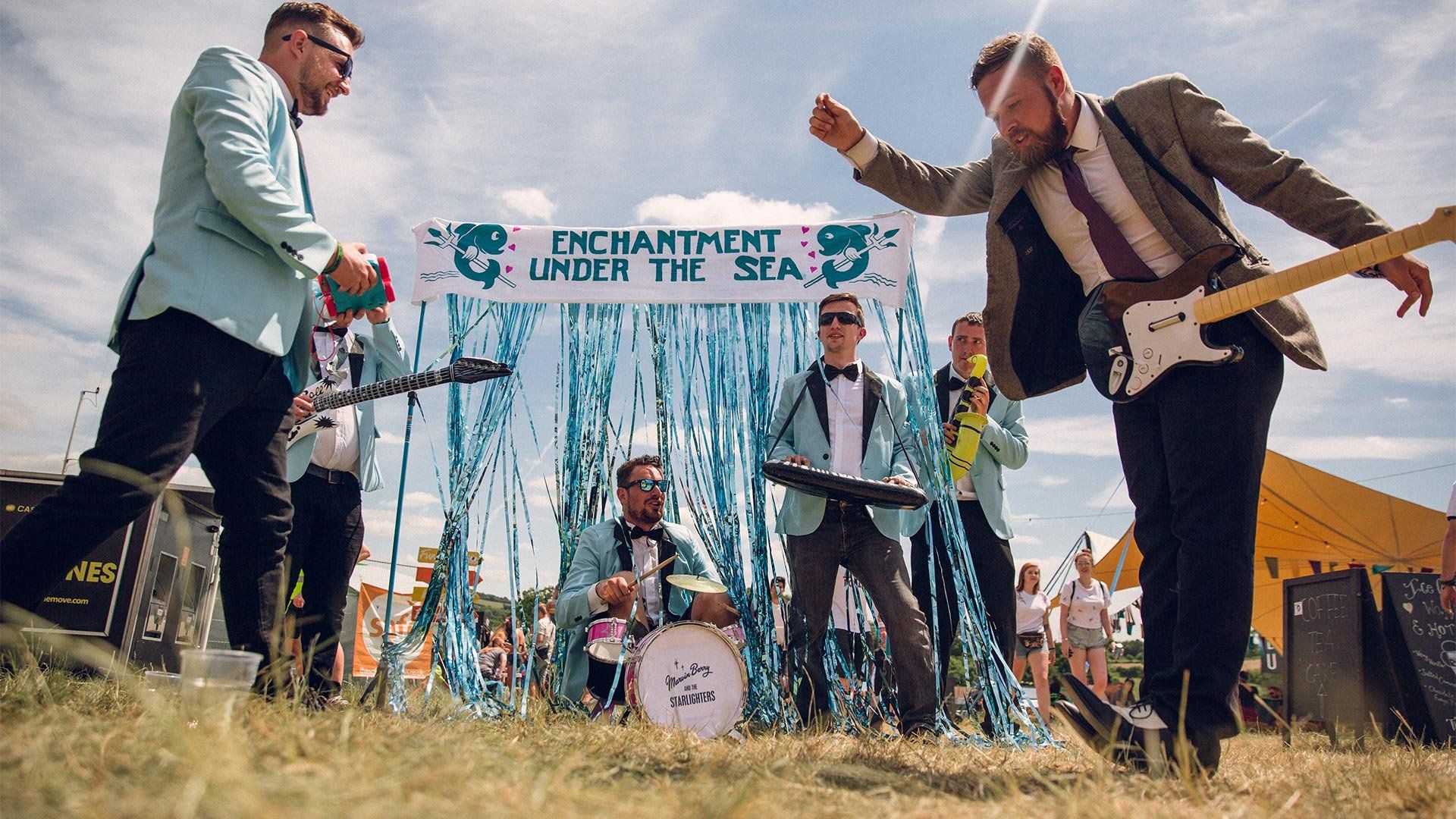 Men in blue jackets and bow ties pretend to play inflatable instruments in a field. Photo by Ben Morse.