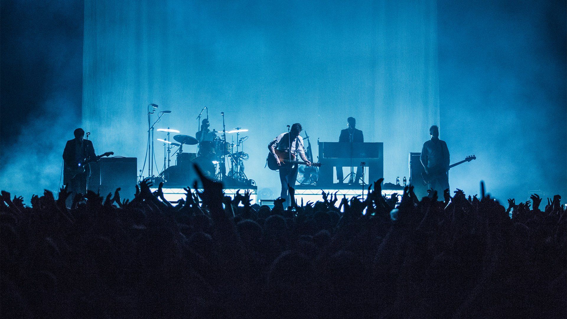A band play on an atmospheric stage at night, with smoke and blue light, and hands in the foreground. Photo by Ben Morse.