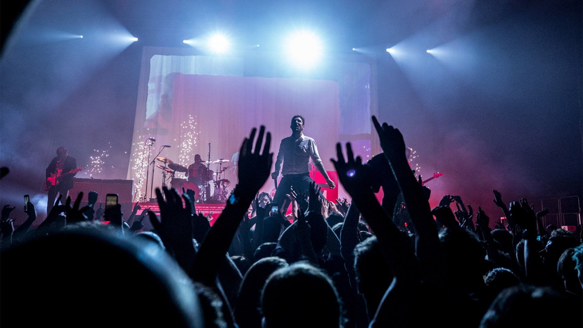 A band perform on stage, with audience hands waving and clapping. Photo by Ben Morse.